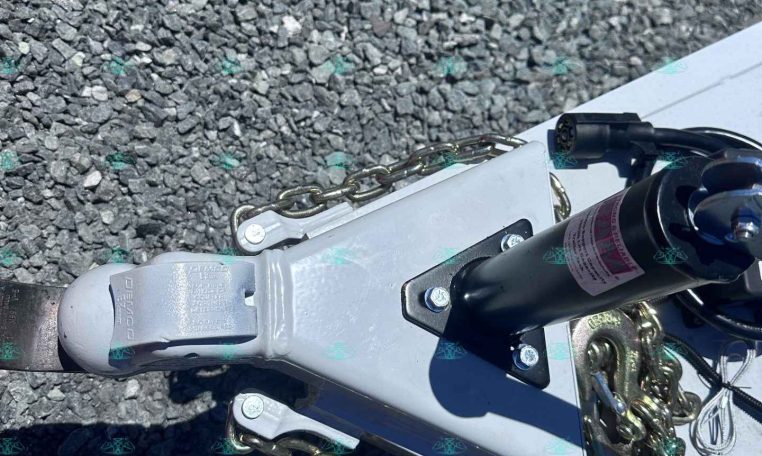 Close-up of a trailer hitch with safety chains attached to a grey metal frame on a gravel surface.