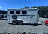 Silver horse trailer with two axles and a mounted spare tire, parked on gravel with a blue sky and trees in the background.