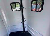 Interior of a light gray horse trailer showing two rounded rectangular windows, a vertical black support post with fold-out brackets, and a dark rubber floor.