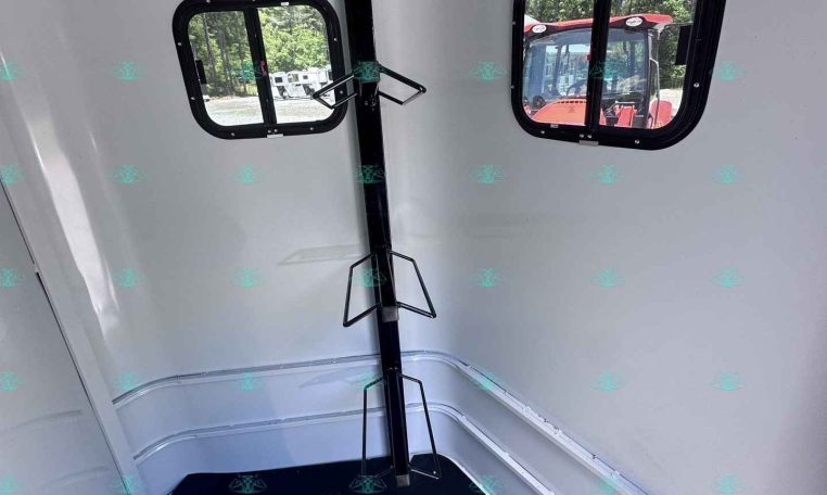 Interior of a light gray horse trailer showing two rounded rectangular windows, a vertical black support post with fold-out brackets, and a dark rubber floor.