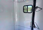Interior of a light gray trailer with a small rounded window and a black vertical support post with a fold-out rack; trees visible outside the window.