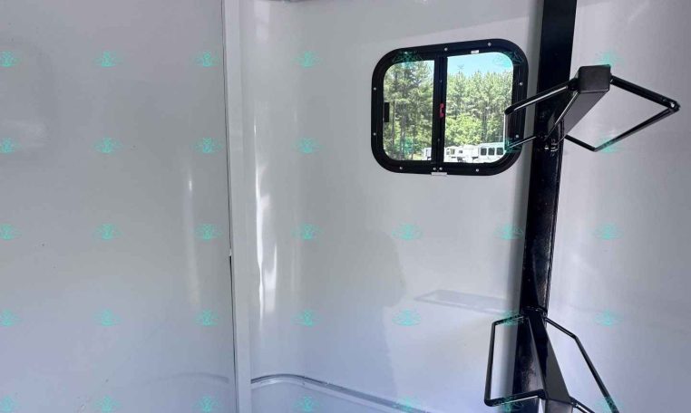 Interior of a light gray trailer with a small rounded window and a black vertical support post with a fold-out rack; trees visible outside the window.
