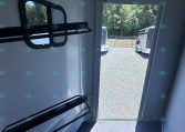 Interior of a light gray trailer with metal wall racks and an open door revealing parked trailers outside on a gravel lot.