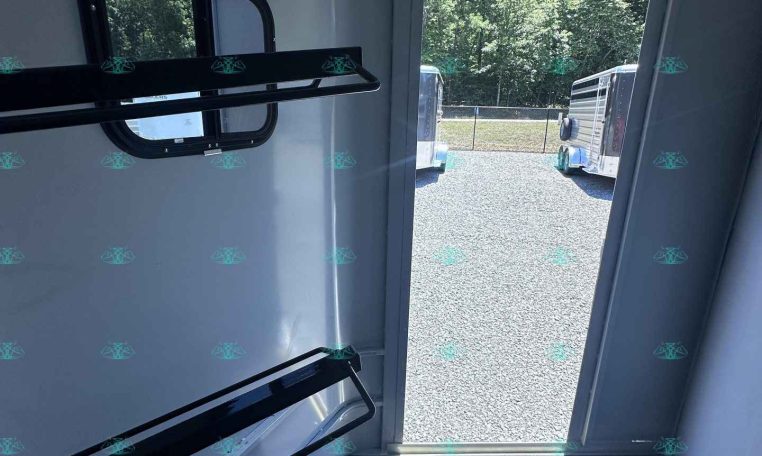 Interior of a light gray trailer with metal wall racks and an open door revealing parked trailers outside on a gravel lot.