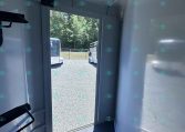 Interior of a light-gray trailer with a doorway open to a gravel lot and parked trailers beyond, trees in the background