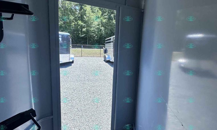 Interior of a light-gray trailer with a doorway open to a gravel lot and parked trailers beyond, trees in the background