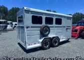 White gooseneck horse trailer with three windows and a side-mounted spare tire, parked on a gravel lot under a clear blue sky.