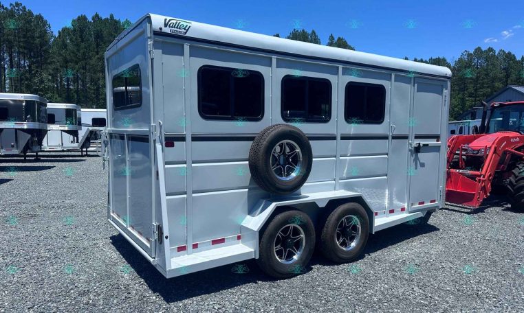 White gooseneck horse trailer with three windows and a side-mounted spare tire, parked on a gravel lot under a clear blue sky.