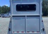 Rear of a white travel trailer with a large rounded window, parked on a gravel lot with trees in the background.