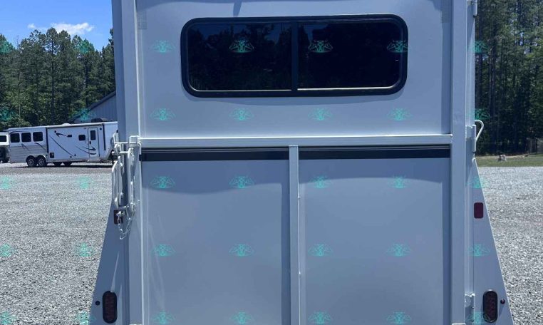 Rear of a white travel trailer with a large rounded window, parked on a gravel lot with trees in the background.