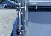 Close-up of a metal trailer hitch latch and safety chains attached to a white trailer, with a gravel lot and a building in the background