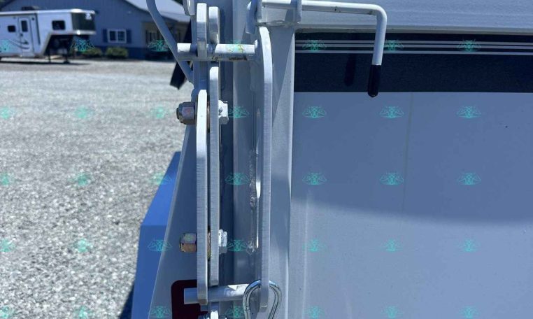Close-up of a metal trailer hitch latch and safety chains attached to a white trailer, with a gravel lot and a building in the background