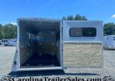 White horse trailer with ramp down, interior showing stalls and wooden walls, parked on a gravel lot with other trailers and trees in the background.