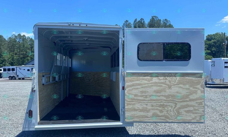 White horse trailer with ramp down, interior showing stalls and wooden walls, parked on a gravel lot with other trailers and trees in the background.