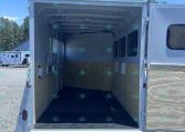 Open cargo trailer with its rear doors down, empty inside, wooden wall panels, and windows along the side, parked on gravel under a blue sky.