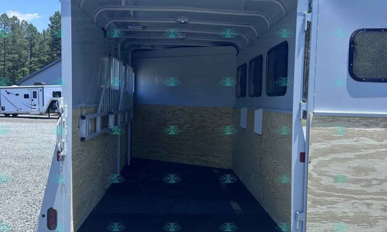 Open cargo trailer with its rear doors down, empty inside, wooden wall panels, and windows along the side, parked on gravel under a blue sky.