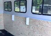 Interior of a light-gray trailer with three black-framed windows along a plywood wall interior, showing outdoors through the glass.