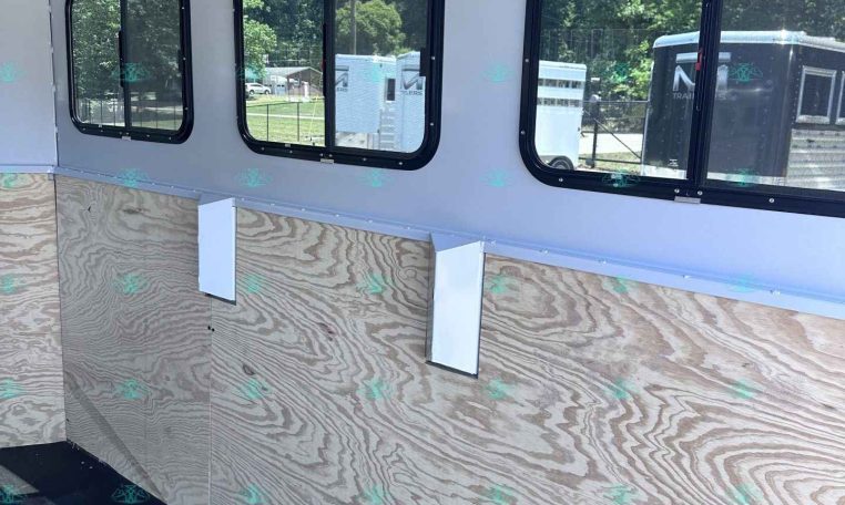 Interior of a light-gray trailer with three black-framed windows along a plywood wall interior, showing outdoors through the glass.