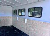 Interior of a light-gray trailer with curved ceiling rails, plywood walls, and several black-framed windows on the side.