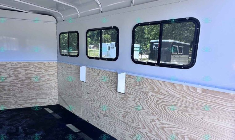 Interior of a light-gray trailer with curved ceiling rails, plywood walls, and several black-framed windows on the side.