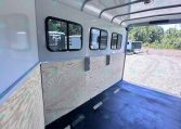 Interior of a light gray trailer with plywood walls, windows along the side, and a dark rubber floor, seen from the open rear doorway.