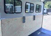 Interior of a light-gray aluminum horse trailer with three rectangular windows and plywood side panels inside. Fixings along the ceiling indicate unfinished interior.