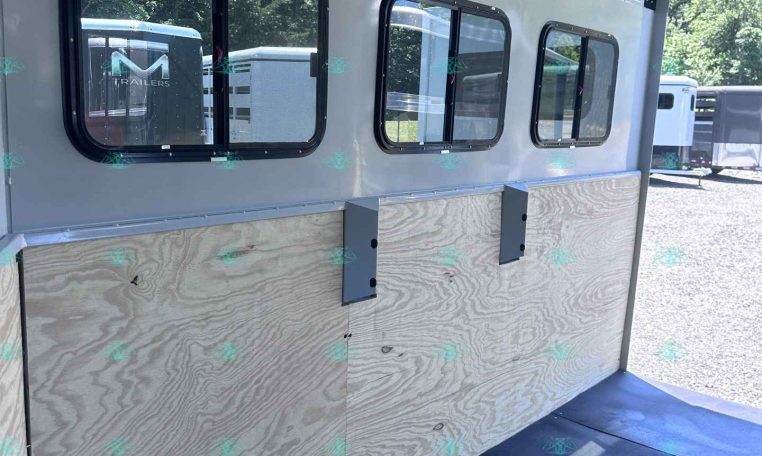 Interior of a light-gray aluminum horse trailer with three rectangular windows and plywood side panels inside. Fixings along the ceiling indicate unfinished interior.