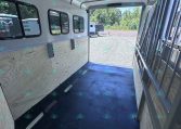 Inside view of a light-gray trailer with plywood sidewalls, small rectangular windows, and a dark rubber floor; a metal divider is on the right and the rear door is open to a gravel lot with other trailers outside.