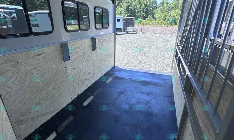 Inside view of a light-gray trailer with plywood sidewalls, small rectangular windows, and a dark rubber floor; a metal divider is on the right and the rear door is open to a gravel lot with other trailers outside.
