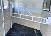 Interior of a white horse trailer with a metal divider and plywood sidewalls, metal stairs/rails on the left, and windows on both sides.