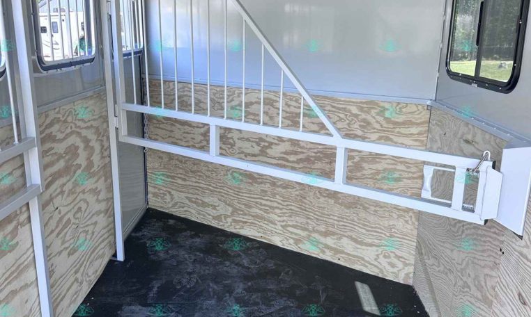 Interior of a white horse trailer with a metal divider and plywood sidewalls, metal stairs/rails on the left, and windows on both sides.