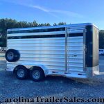 Silver aluminum, two-axle horse trailer parked on a gravel lot with trees in the background and a building nearby.