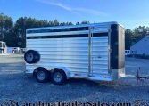 Silver aluminum, two-axle horse trailer parked on a gravel lot with trees in the background and a building nearby.