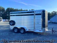 Silver aluminum, two-axle horse trailer parked on a gravel lot with trees in the background and a building nearby.