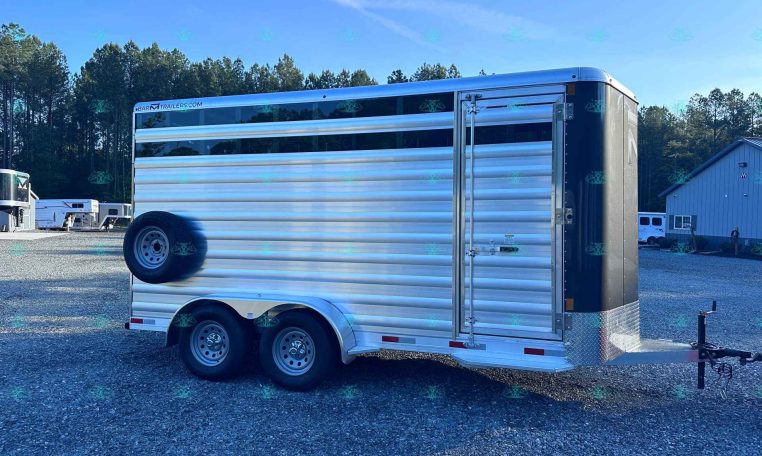 Silver aluminum, two-axle horse trailer parked on a gravel lot with trees in the background and a building nearby.