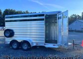 Aluminum two-axle livestock trailer with open rear double doors, parked on gravel with a watermarked dealership site visible.