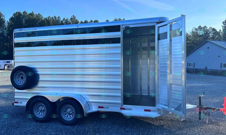 Aluminum two-axle livestock trailer with open rear double doors, parked on gravel with a watermarked dealership site visible.
