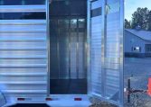 Silver aluminum trailer with its rear door wide open, revealing a bare interior with vertical dividers, parked on a gravel lot under a blue sky.