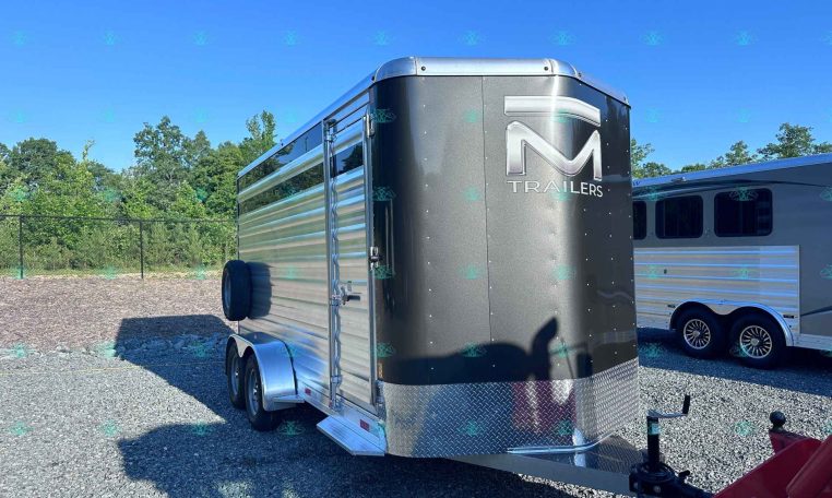Shiny silver cargo trailer with diamond‑plate front and 'M Trailers' logo, parked on a gravel lot with a blue sky.