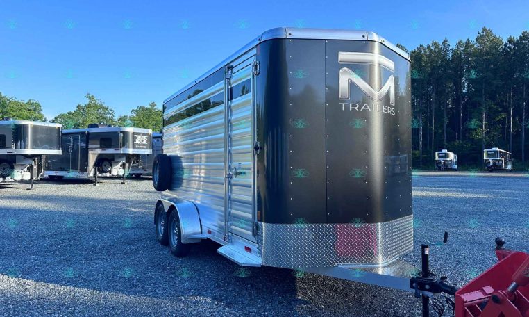 Large black enclosed horse trailer with chrome diamond-plate front, parked in a gravel lot among other trailers, trees in the background.