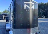 Gray enclosed trailer with a large silver 'M' logo and the word TRAILERS on the back, chrome trim, and a diamond-plate front, parked on a gravel lot under a clear blue sky with trees in the background.
