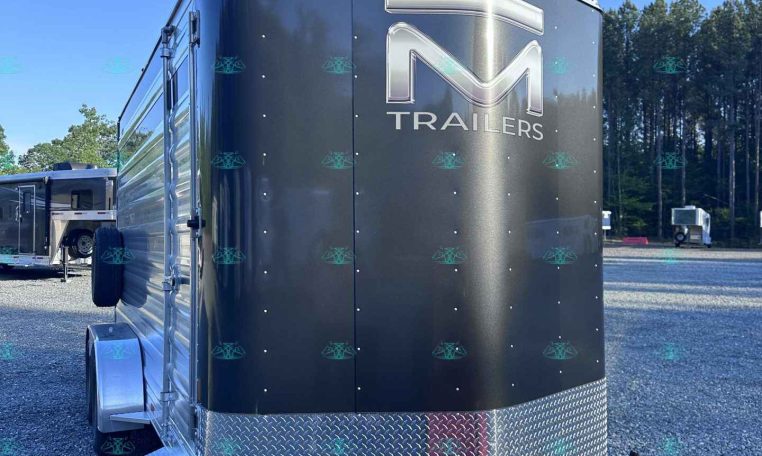 Gray enclosed trailer with a large silver 'M' logo and the word TRAILERS on the back, chrome trim, and a diamond-plate front, parked on a gravel lot under a clear blue sky with trees in the background.