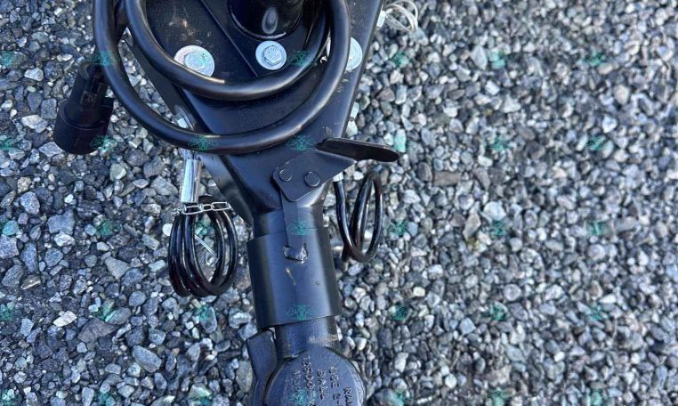 Close-up of a trailer hitch coupler and wiring harness on a metal frame, set on gravel ground.