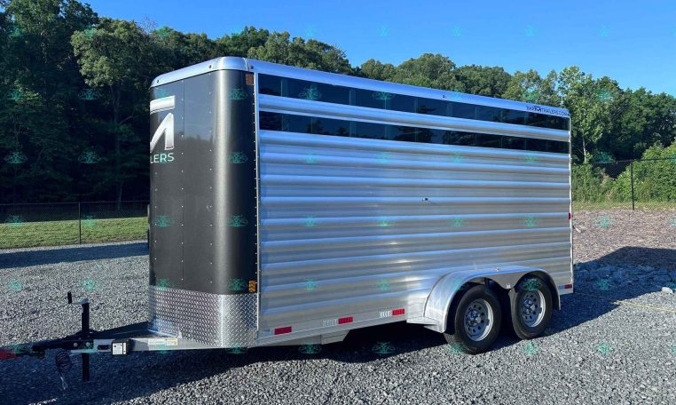Side view of a silver enclosed cargo trailer on a gravel lot with trees and a clear blue sky behind it.