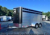 Silver and black aluminum livestock trailer with dual axles parked in a lot, with other trailers and green trees in the background under a clear blue sky