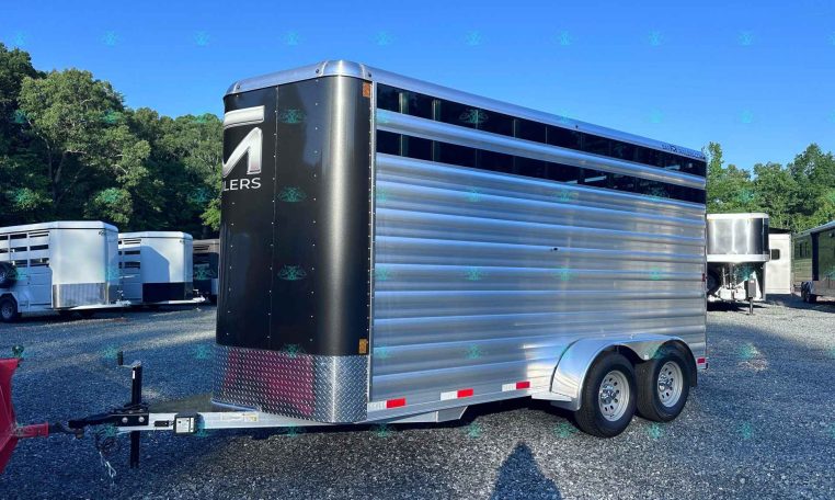 Silver and black aluminum livestock trailer with dual axles parked in a lot, with other trailers and green trees in the background under a clear blue sky