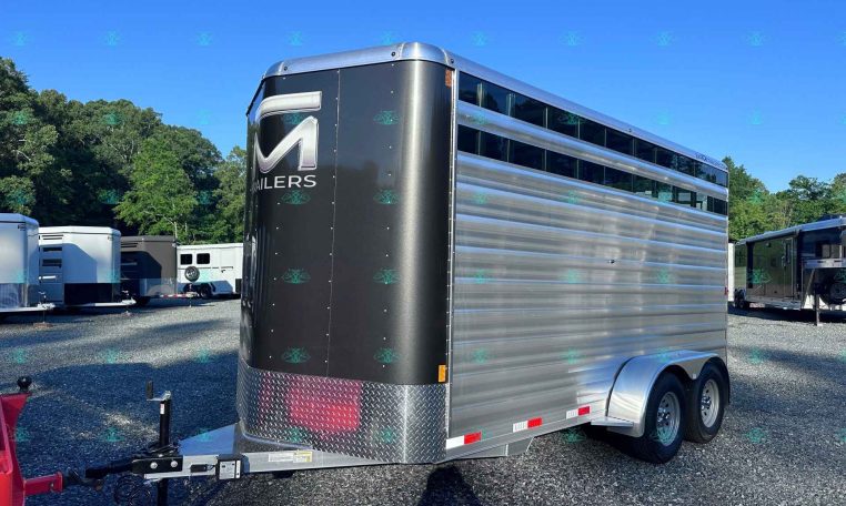 Large aluminum trailer with a black front panel and chrome diamond-plate lower edge, parked with other trailers on a gravel lot under a blue sky.