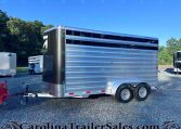 Silver aluminum horse trailer with two axles and black front, parked on a gravel lot with other trailers, blue sky and trees in the background, CarolinaTrailersales.com watermark at bottom.