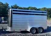 Shiny aluminum livestock trailer with black front corner, parked on a gravel lot with trees and blue sky behind.