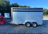 Silver aluminum livestock trailer parked on a gravel lot with a row of trees in the background.